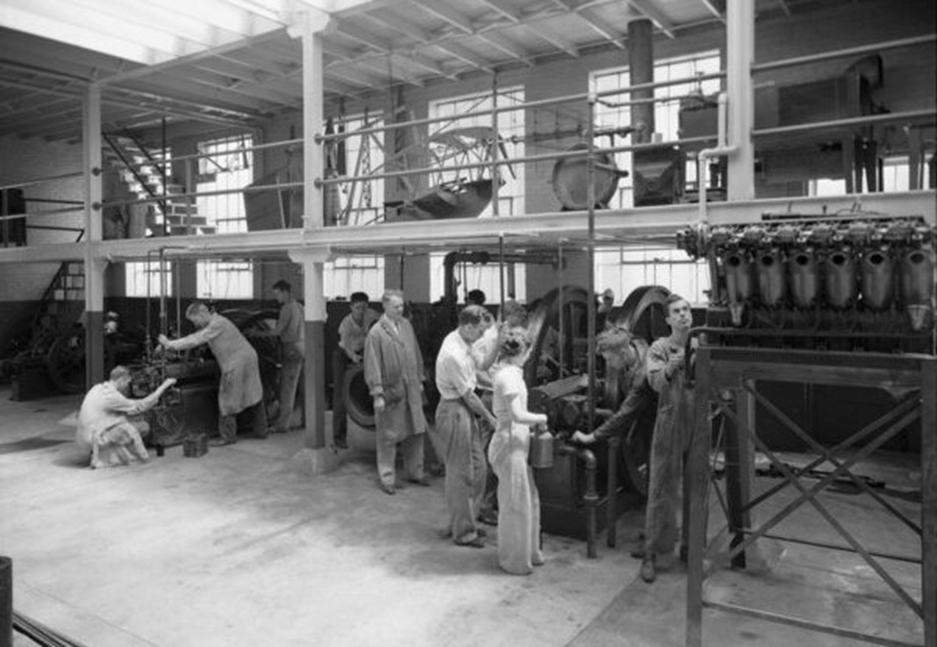 Black and white photo of male engineering students surrounding the singular female engineering student during a co-op at a factory. August 1936.