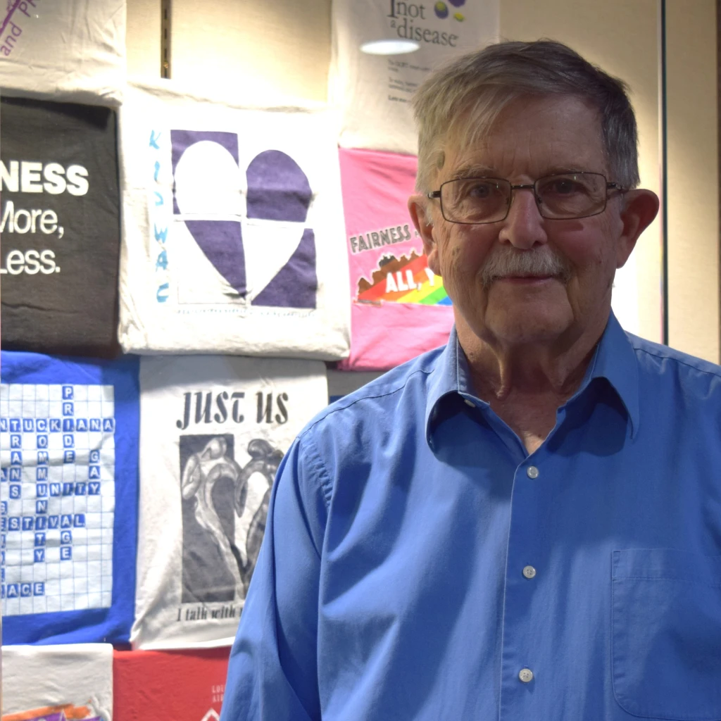 David Williams stands in front of a colorful display of t-shirts from various LGBTQ+ movements across Kentucky.